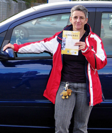 Frau Benz steht vor ihrem Auto und zeigt die gewonnene Urkunde in die Kamera