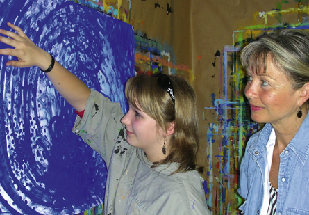 Erika Syska-Reis schaut einer jungen Malerin über die Schulter, während diese einen blauen Wirbel auf die Leinwand bringt