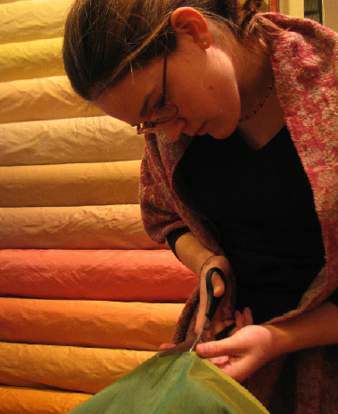 junge Dame beim Zuschneiden grüner Seide - im Hintergrund gestapelte Seide in Tönen von gelb bis rot 
